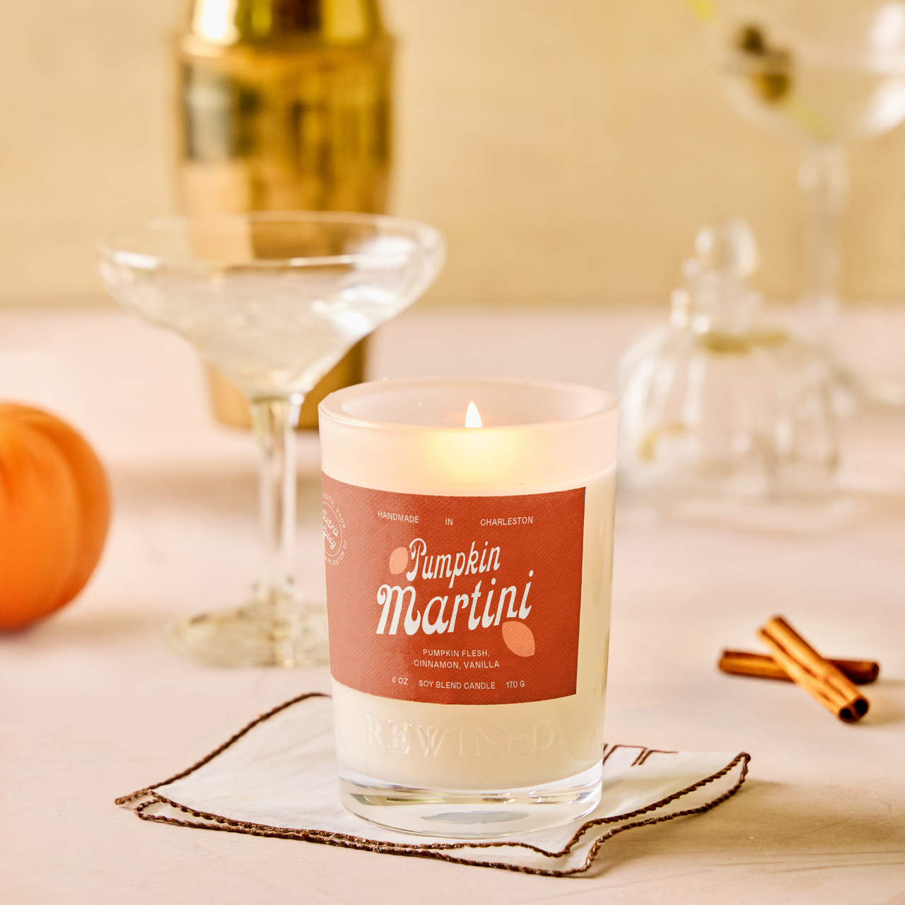 A lit scented candle in a clear glass jar sits on a napkin on a tabletop, with a warm autumn-themed background including a small orange pumpkin, cinnamon sticks, and cocktail glasses. The candle label reads 'Pumpkin Martini'.